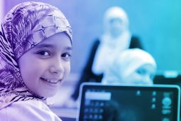 طفلة صغيرة مبتسمة و الكمبيوتر المحمول مفتوح أمامها و المعلمة في الخلفية تتكلم مع الطلاب.