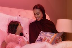 A mother wearing traditional Qatari clothes, is sitting in bed next to her little daughter and reading her a bedtime story.