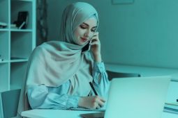  امرأة ترتدي الحجاب ، وتكتب على مفكرة بينما تتكلم على هاتفهها الذكي وحاسوبها المحمول مفتوح أمامها.