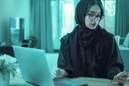  امرأة ترتدي الحجاب وتتفحص هاتفها الذكي وتستخدم جهاز الكمبيوتر المحمول الخاص بها.