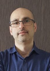 Raian Ali wearing a dark blue shirt and eyeglasses. The background is a brown wall.